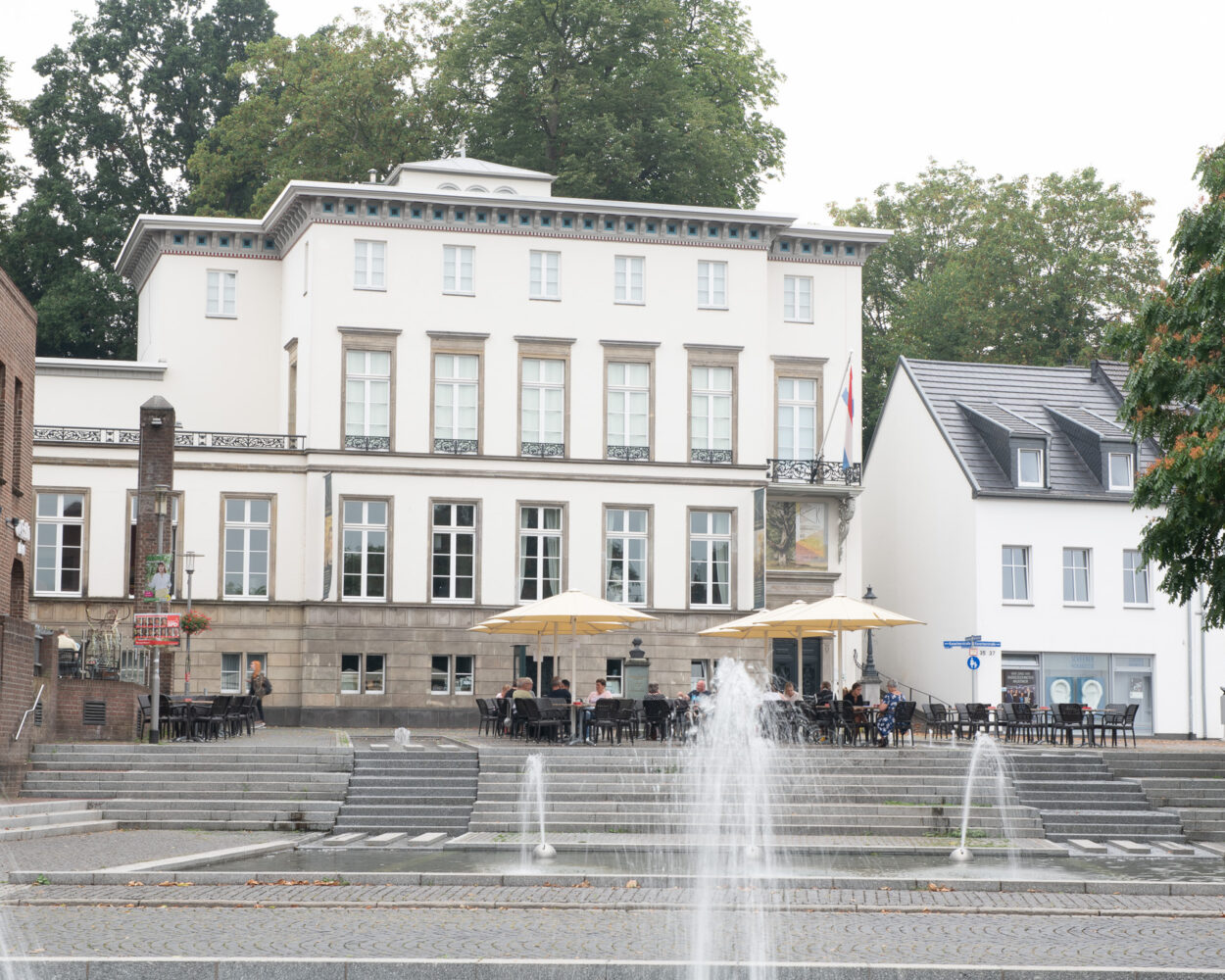 Mehrstöckiges weißes Gebäude des Museum B.C. Koekkoek-Haus mit vielen Fenstern und zwei großen Sonnenschirmen vor der Fassade, davor Treppen und mehrere Wasserfontänen.