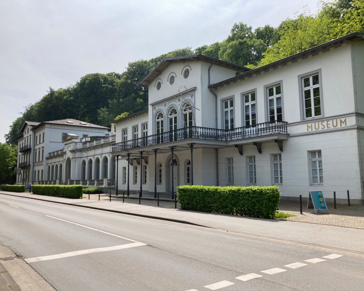 Fassade des Museum Kurhaus Kleve mit weißen Wänden, großen Fenstern und einem Balkon. Im Vordergrund eine Straße mit Bäumen und einem Informationsschild.