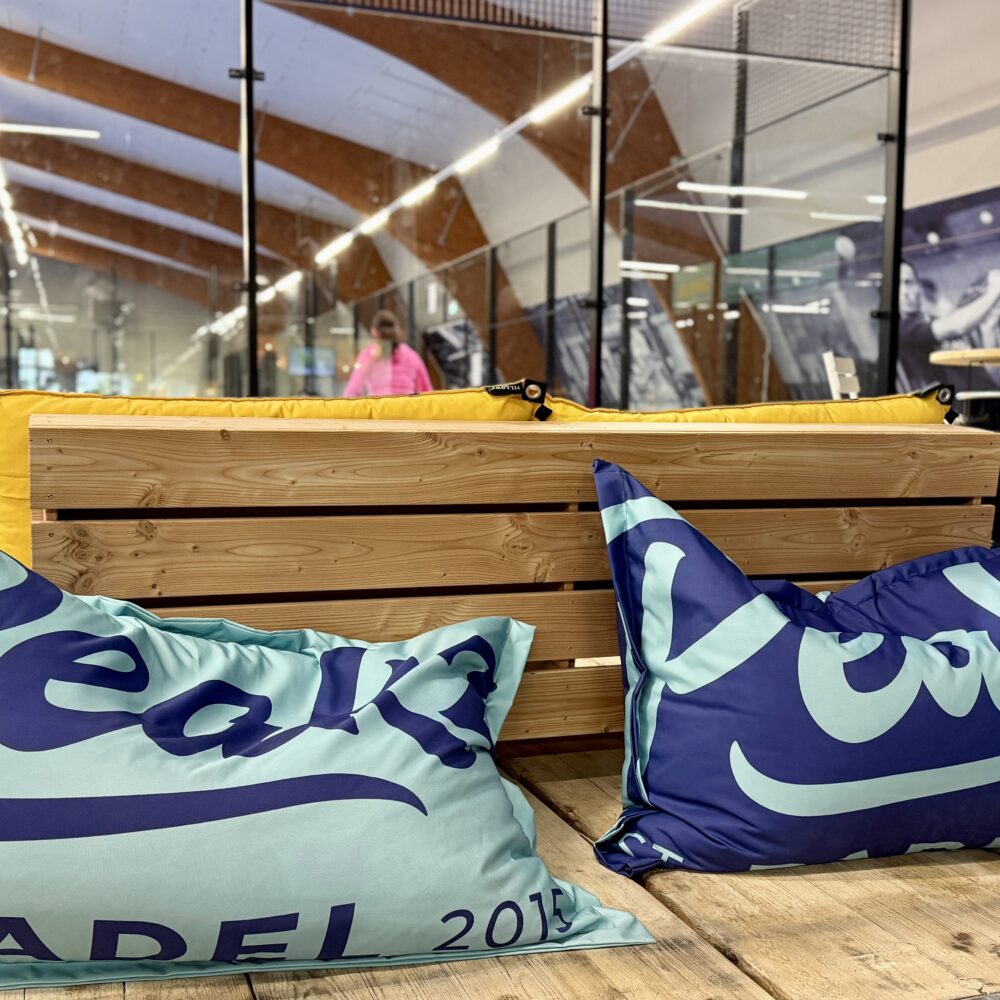 Zwei große Kissen mit dem Aufdruck 'Peakz Padel 2019' auf einer Holzplattform. Im Hintergrund sind Padelplätze und eine Person sichtbar.
