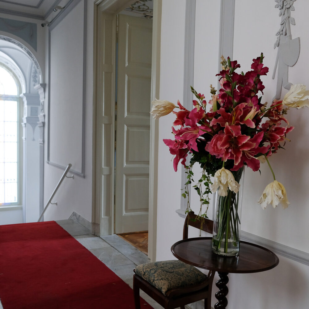Flur im Museum B.C. Koekkoek-Haus mit rotem Teppich, großem Fenster mit Rundbogen und Vase mit roten und weißen Blumen auf einem runden Holztisch neben zwei Stühlen.