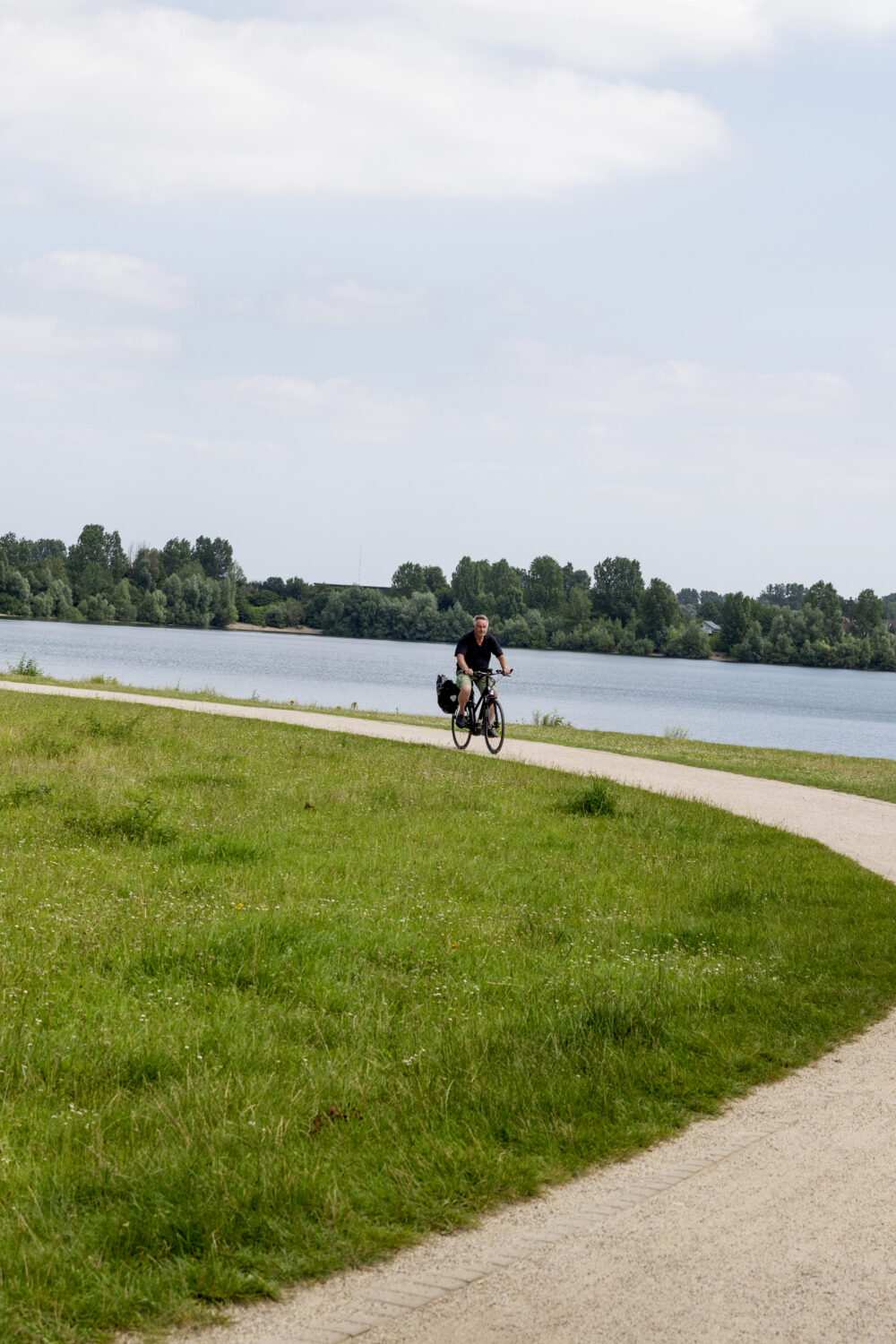 Radfahrer auf geschwungenem Weg am Rheinufer, daneben zwei Spaziergänger auf Grasfläche unter bewölktem Himmel.