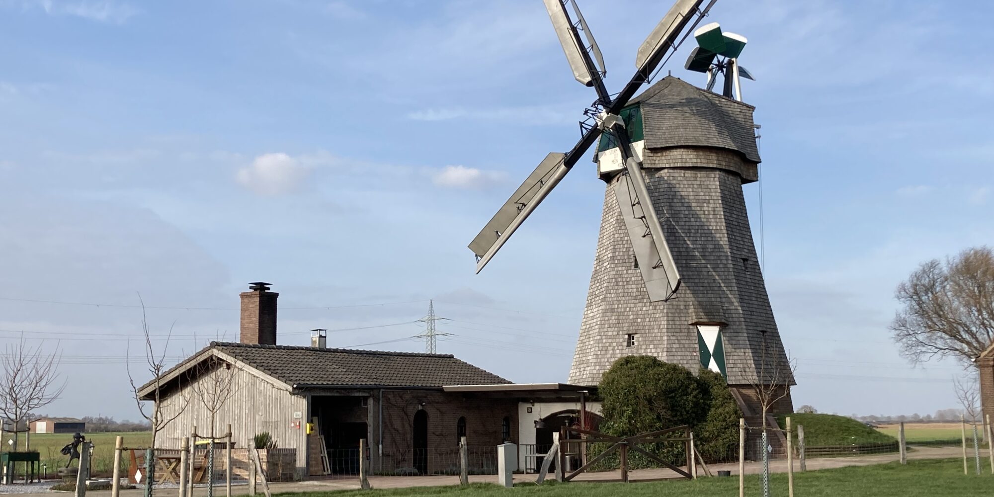Eine alte Mühle mit grauem Mauerwerk und großen, weißen Flügeln steht in einer grünen Wiese. Neben der Mühle befindet sich ein Nebengebäude aus Holz.