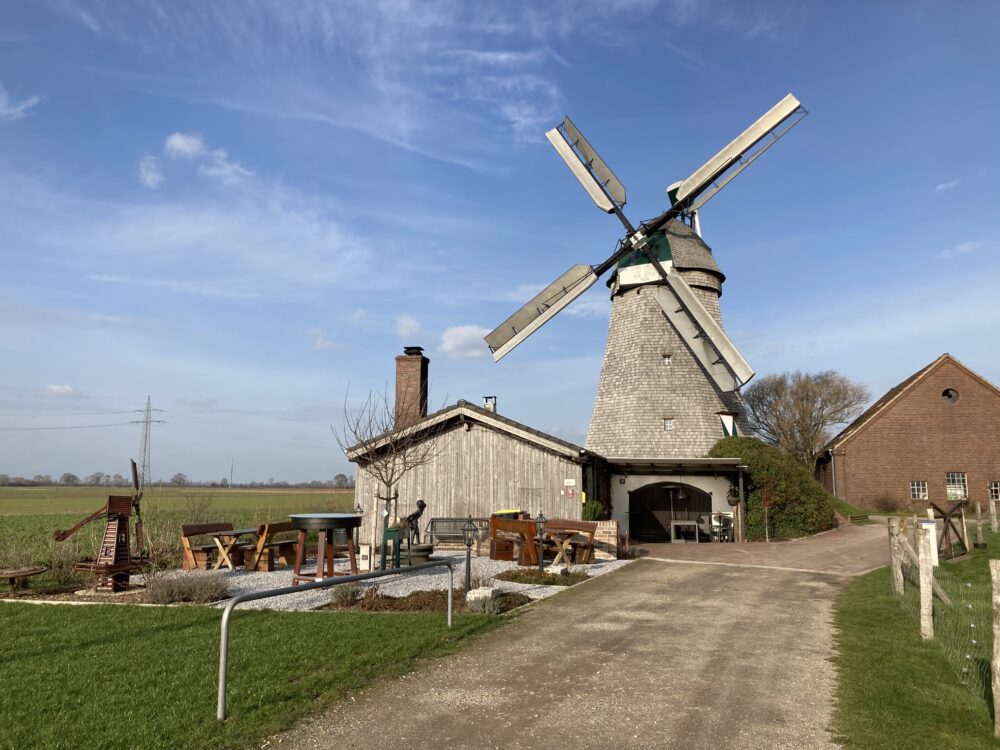 Eine alte Mühle mit grauem Steingebäude und vier großen, beweglichen Flügeln. Im Vordergrund sind Gartenmöbel und eine Statue sichtbar.