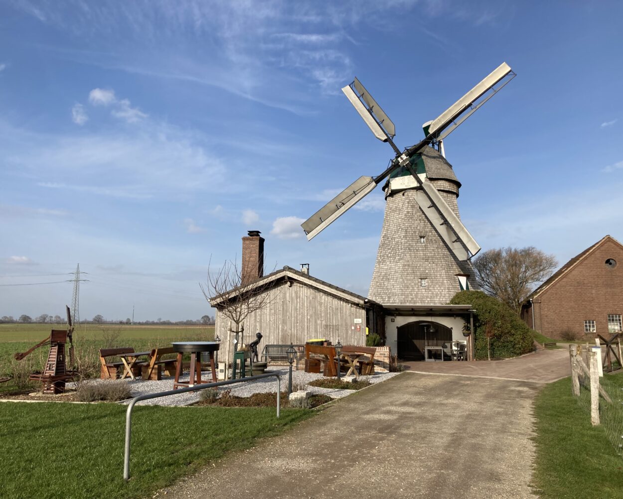 Eine alte Mühle mit grauem Steingebäude und vier großen, beweglichen Flügeln. Im Vordergrund sind Gartenmöbel und eine Statue sichtbar.