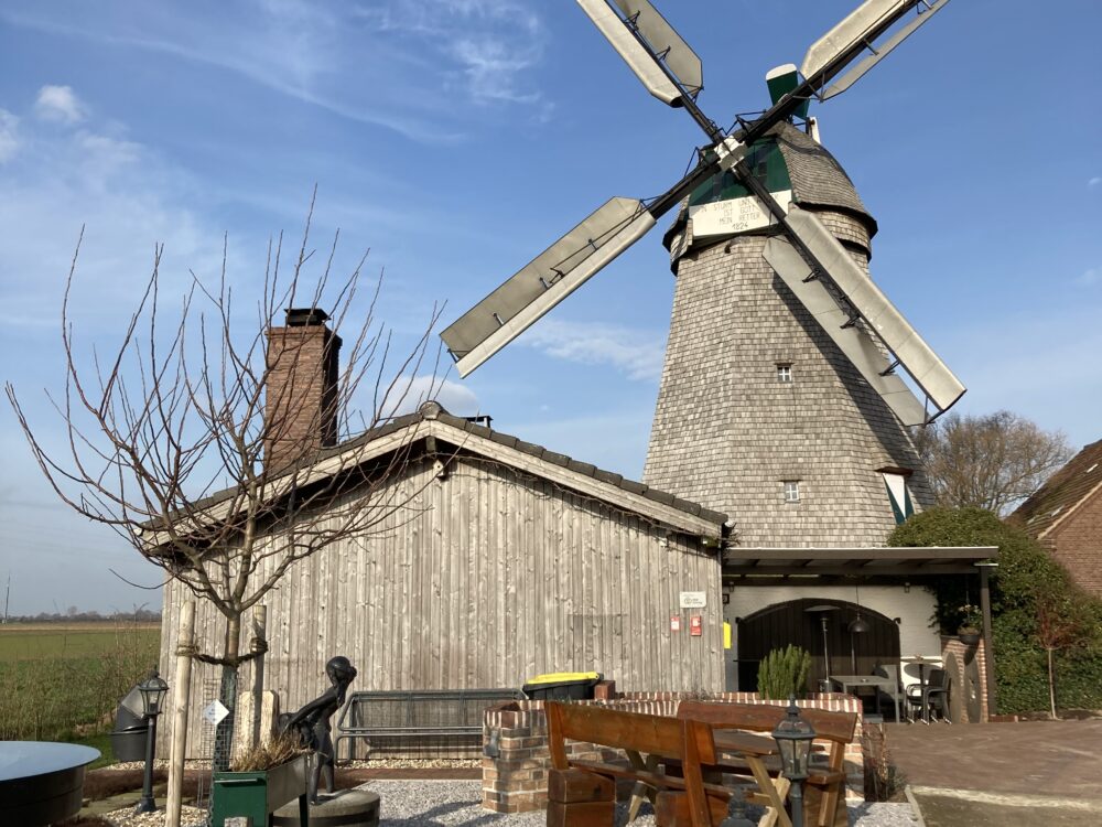 Eine alte Mühle mit grauem Steingebäude und vier großen, beweglichen Flügeln. Im Vordergrund sind Gartenmöbel und eine Statue sichtbar.