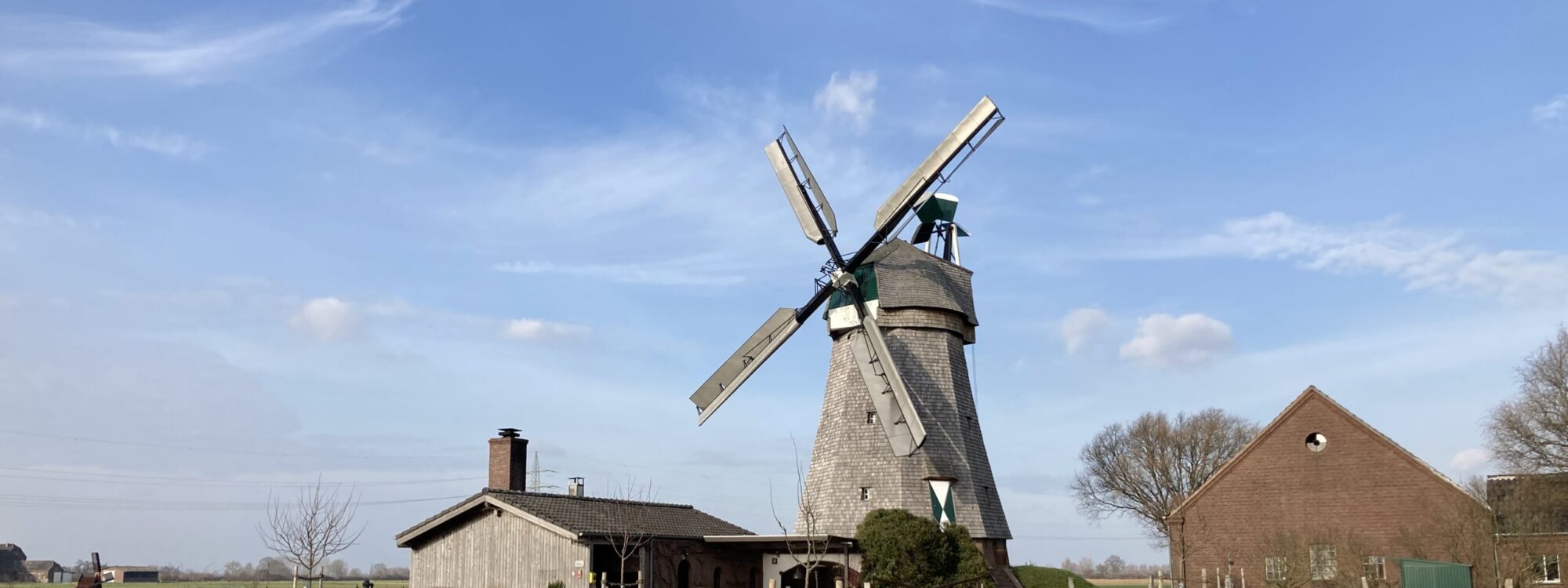 Eine alte Mühle mit grauem Mauerwerk und großen, weißen Flügeln steht in einer grünen Wiese. Neben der Mühle befindet sich ein Nebengebäude aus Holz.