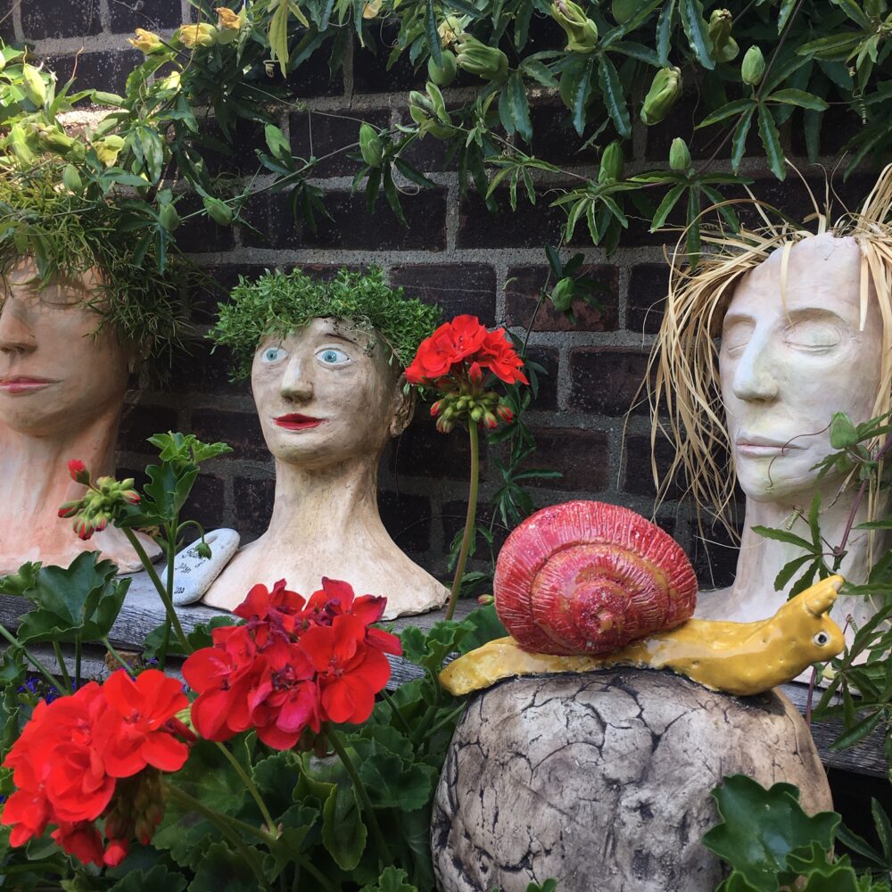 Rote Geranienblüten vor drei weißen Keramikgesichtern und einer gelben Schnecke mit rotem Gehäuse auf einem Stein vor einer Backsteinmauer mit grünem Efeu.