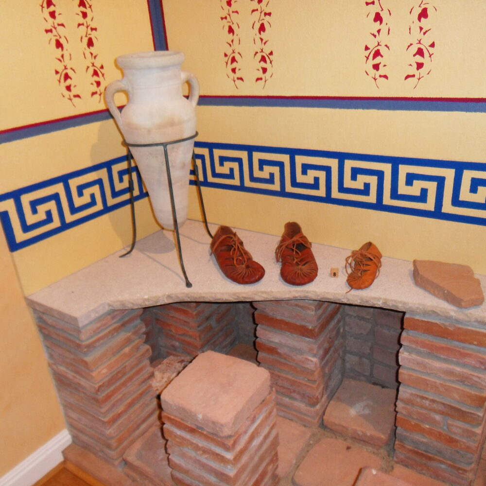 Nachbildung einer römischen Fußbodenheizung aus alten Ziegelsteinen und einer Bodenplatte. Darauf römische Schuhe und eine Vase. An der Wand Malereien