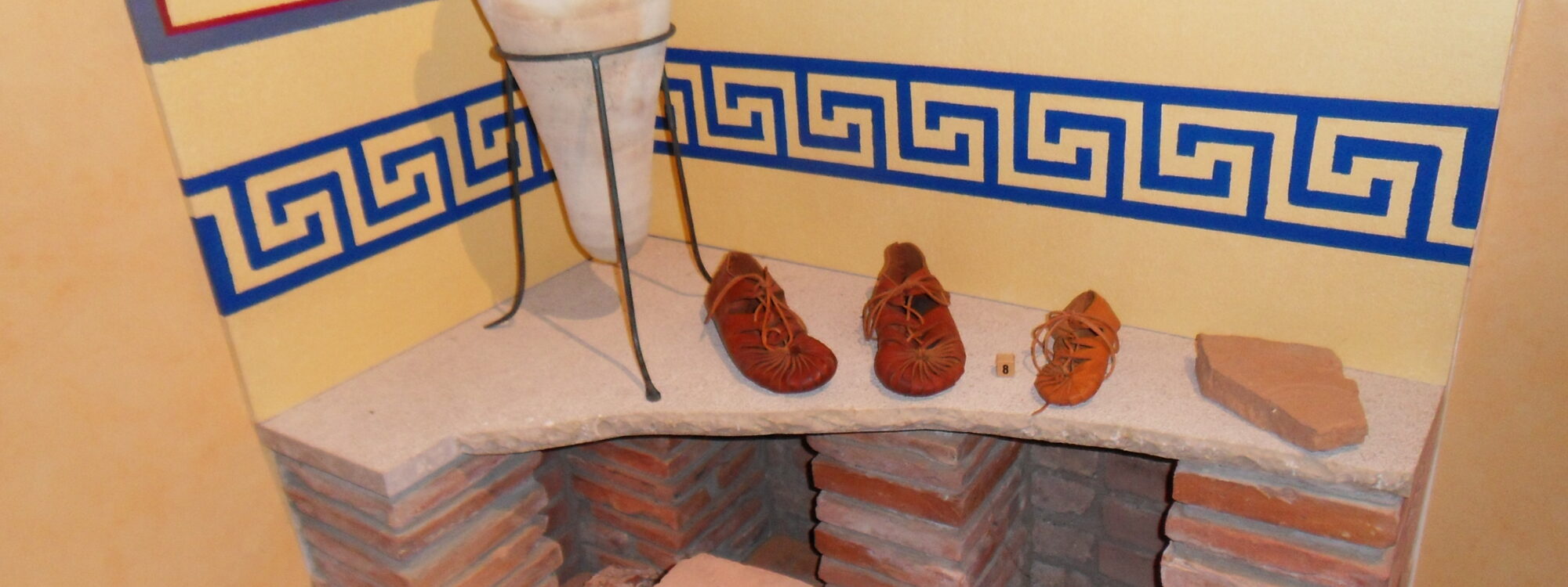 Nachbildung einer römischen Fußbodenheizung aus alten Ziegelsteinen und einer Bodenplatte. Darauf römische Schuhe und eine Vase. An der Wand Malereien