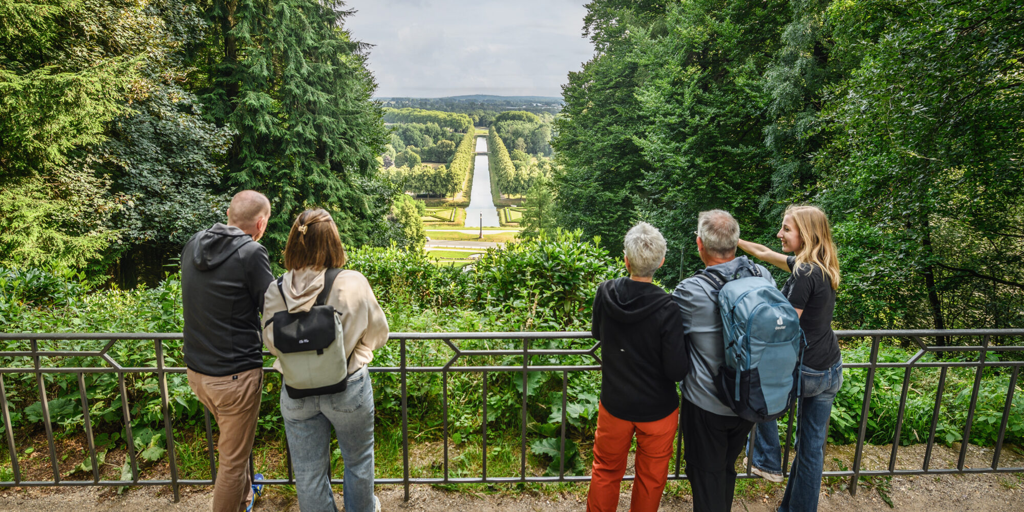 Fünf Personen stehen an einem Geländer und blicken auf eine weitläufige Parklandschaft mit einem Wasserlauf in der Mitte.