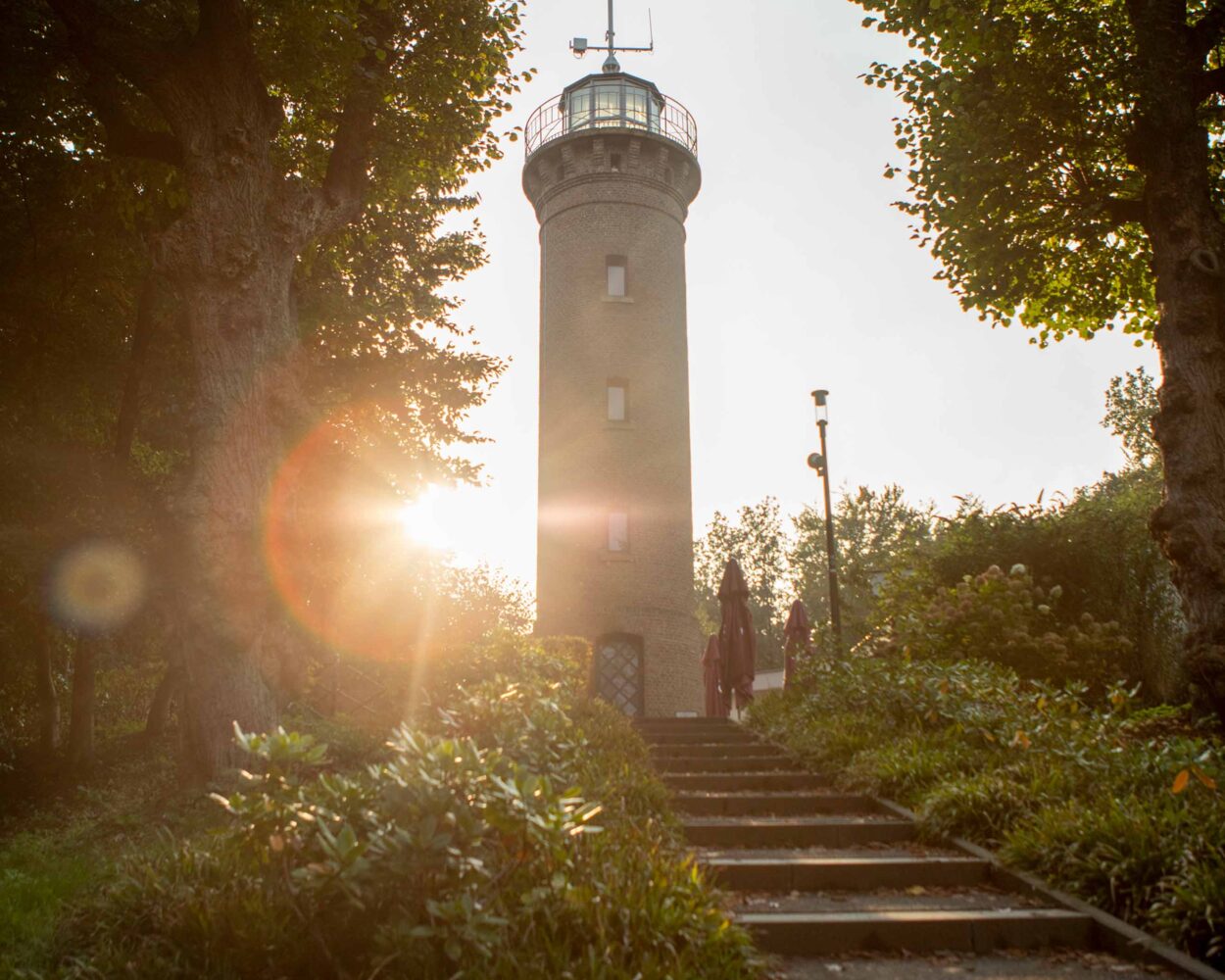 Ein Aussichtsturm aus Ziegelstein mit einem runden oberen Teil und Antennen, umgeben von großen, runden Bäumen. Im Hintergrund ist ein bewölkter Himmel zu sehen.