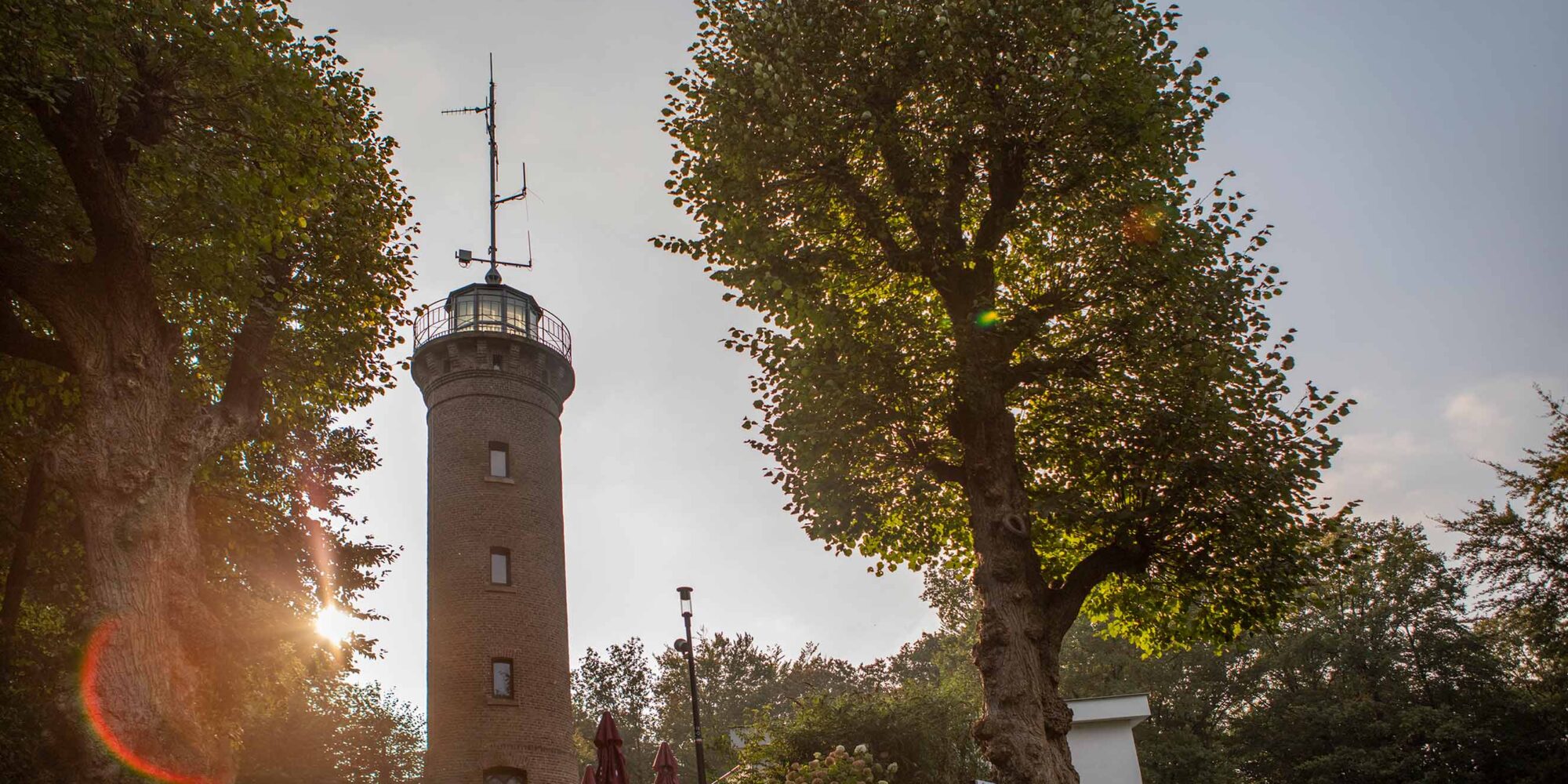 Ein Aussichtsturm aus Ziegelstein mit einem runden oberen Teil und Antennen, umgeben von großen, runden Bäumen. Im Hintergrund ist ein bewölkter Himmel zu sehen.