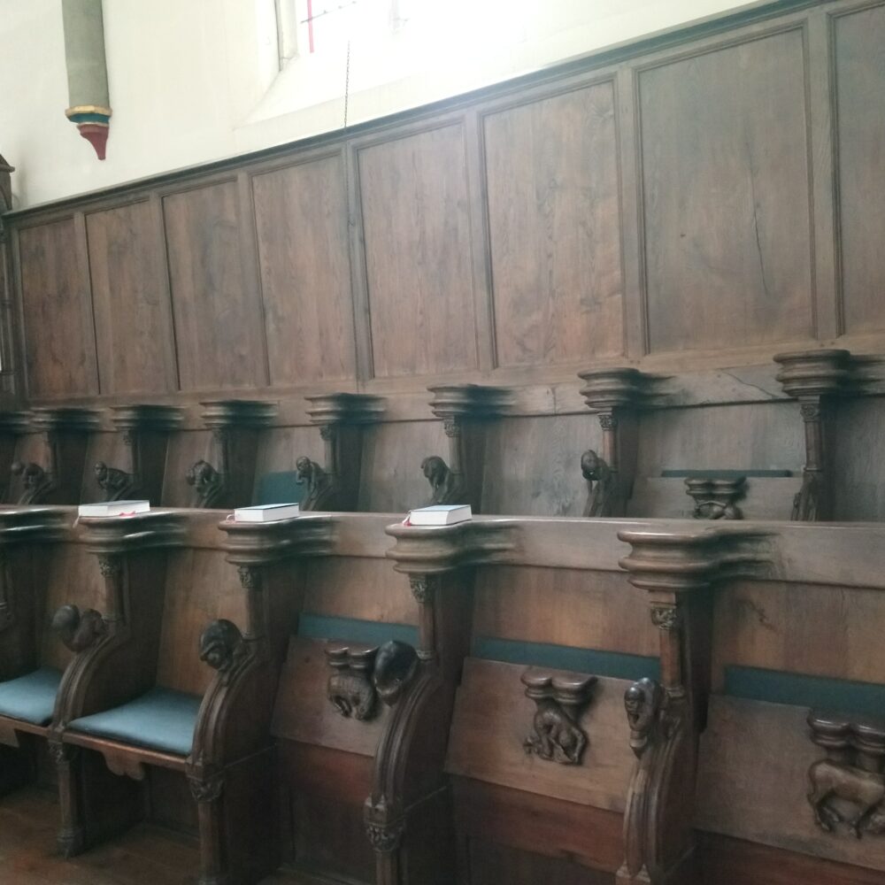 Holz-Chorgestühl in der Minoritenkirche mit geschnitzten Verzierungen und gepolsterten Sitzen. Es sind insgesamt acht Sitzplätze sichtbar.