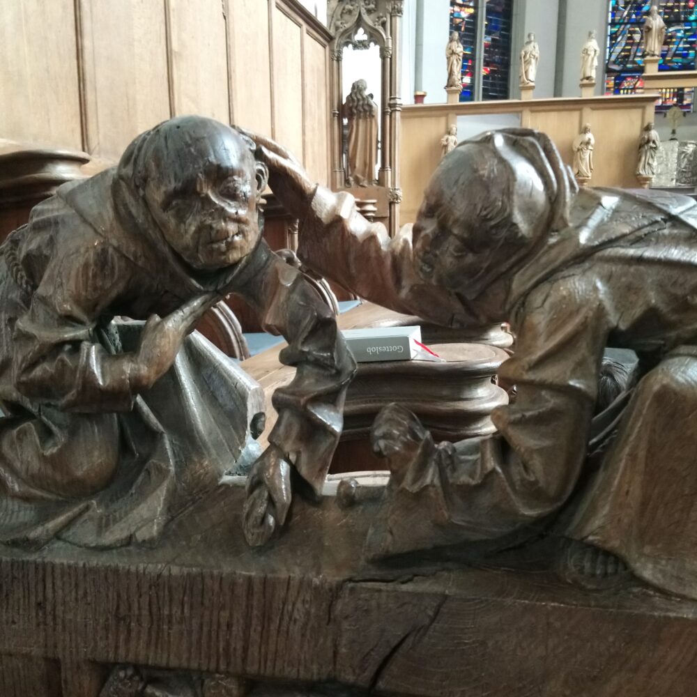 Zwei geschnitzte Figuren in einem Chorgestühl, eine Figur berührt die andere am Kopf. Im Hintergrund sind farbige Fenster und Statuen sichtbar.