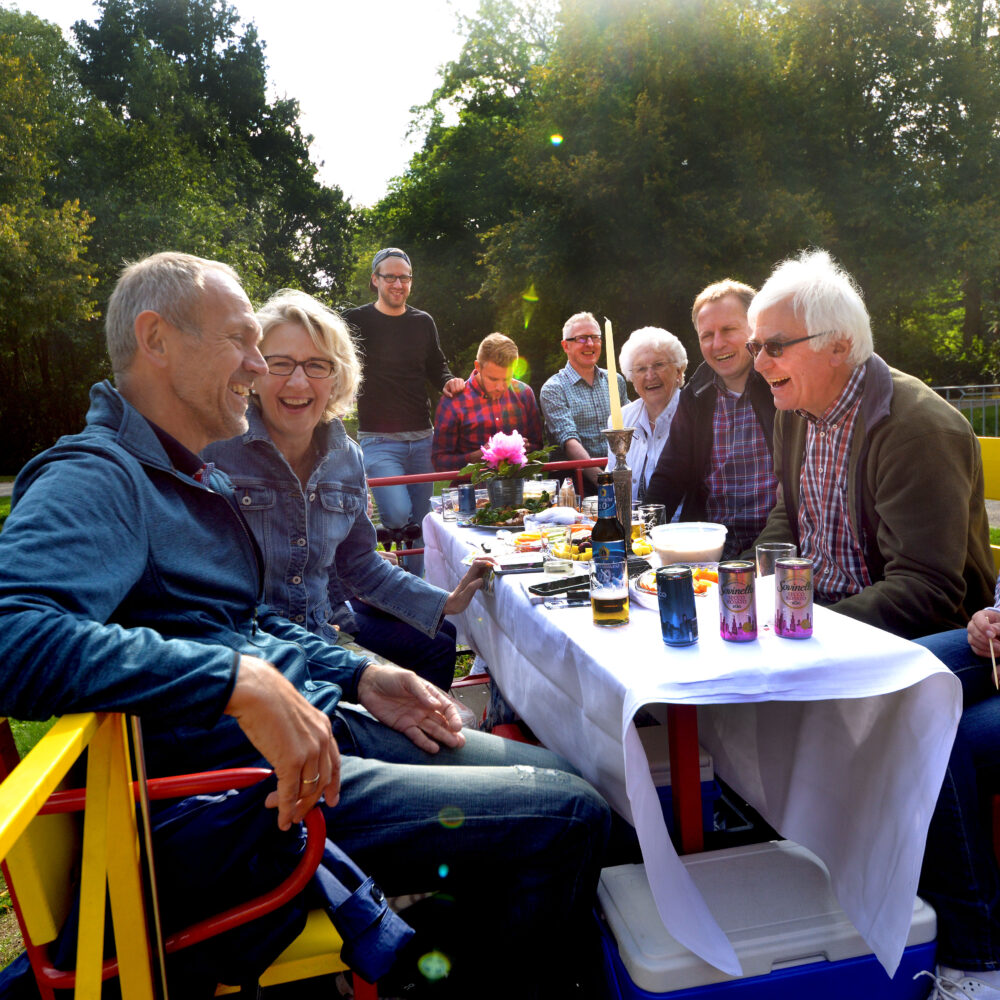 Eine Gruppe von Personen sitzt an einem Tisch mit einem weißen Tischtuch auf einer Clubdraisine. Auf dem Tisch stehen Getränke und Snacks. Im Hintergrund sind Bäume und ein Weg sichtbar.