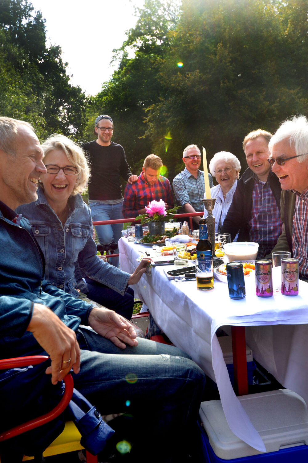 Eine Gruppe von Personen sitzt an einem Tisch mit einem weißen Tischtuch auf einer Clubdraisine. Auf dem Tisch stehen Getränke und Snacks. Im Hintergrund sind Bäume und ein Weg sichtbar.