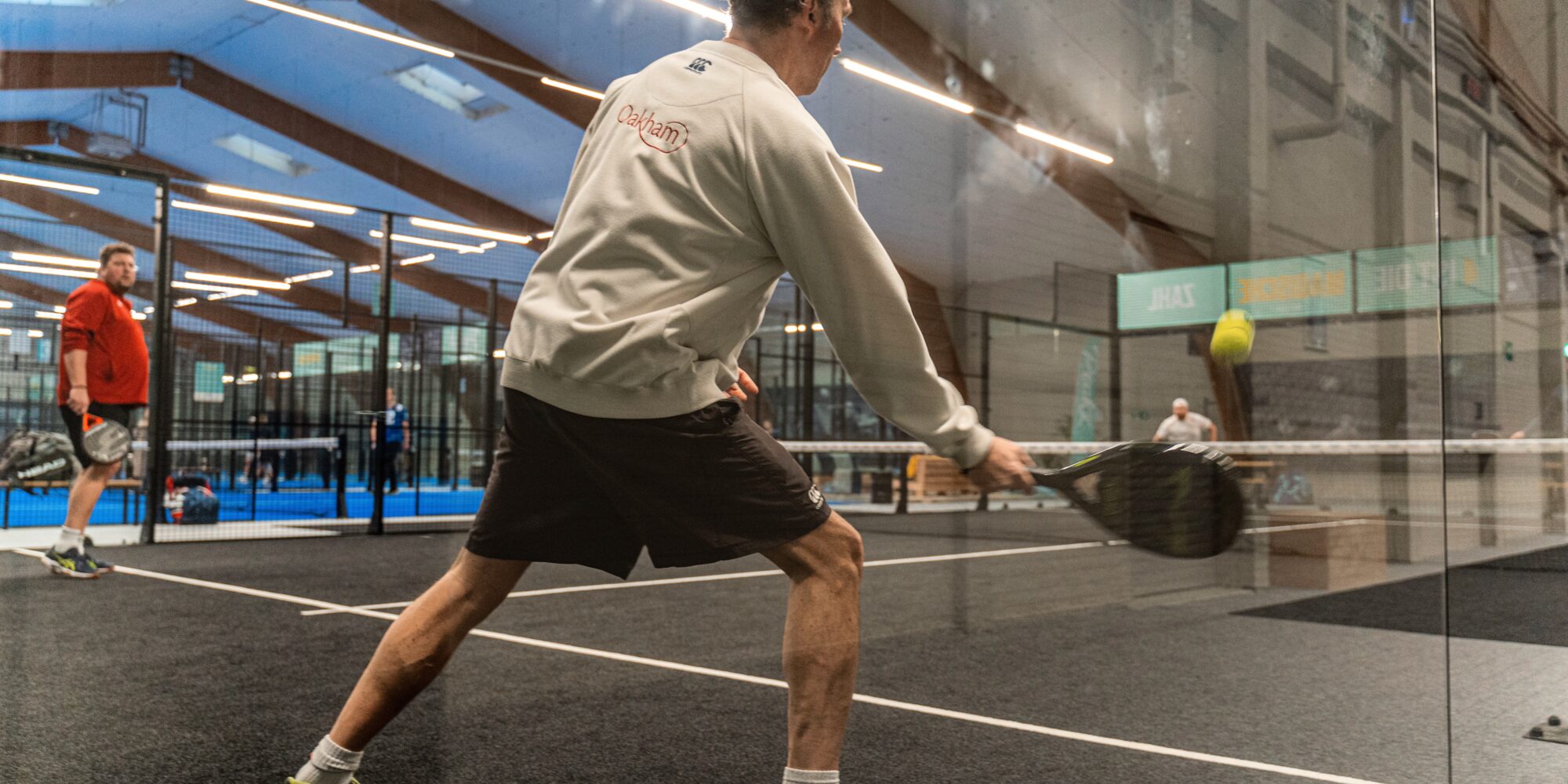 Ein Mann in einem grauen Sweatshirt und schwarzen Shorts schlägt mit einem Schläger einen Tennisball. Im Hintergrund sind weitere Spieler und ein Sportplatz sichtbar.