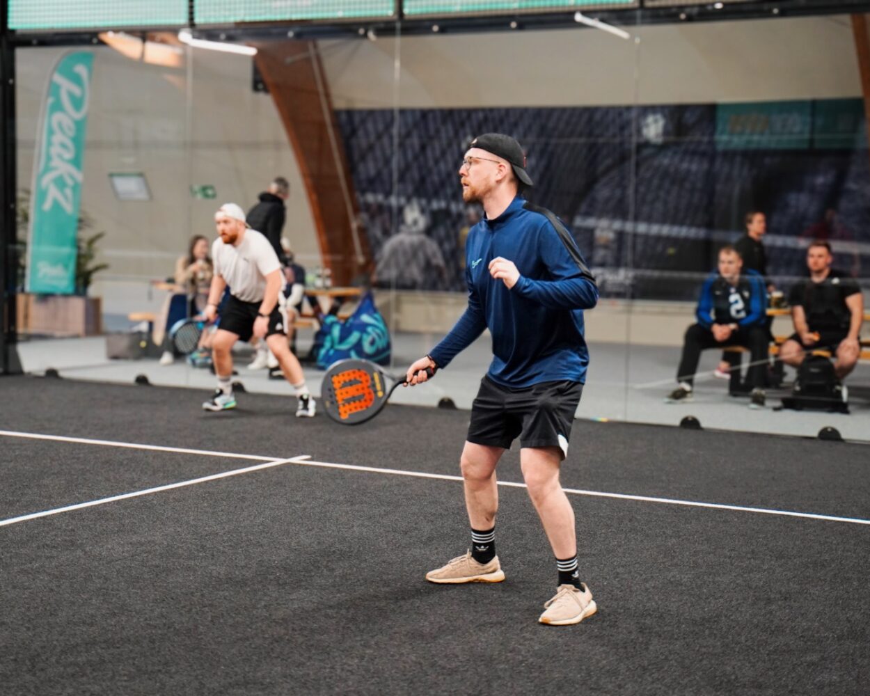 Zwei Personen spielen Padel-Tennis in einer Halle. Eine Person in blauer Jacke und schwarzer Shorts steht bereit, während die andere Person im Hintergrund einen Schlag ausführt.