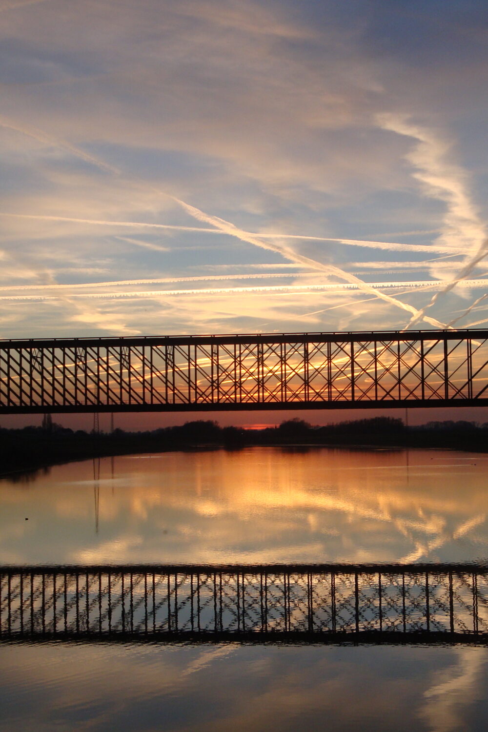 Die alte Bahnbrücke Griethausen ist eine hohe, metallene Fachwerkbrücke, die sich über einen ruhigen Fluss spannt. Ihre Silhouette spiegelt sich im Wasser.