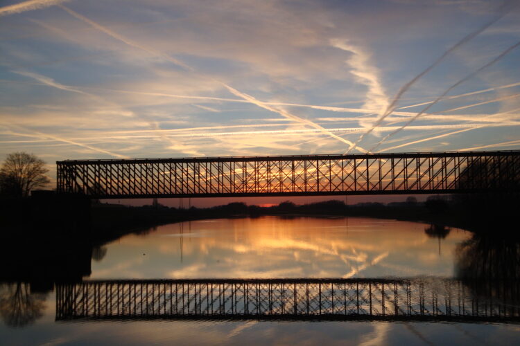 Die alte Bahnbrücke Griethausen ist eine hohe, metallene Fachwerkbrücke, die sich über einen ruhigen Fluss spannt. Ihre Silhouette spiegelt sich im Wasser.
