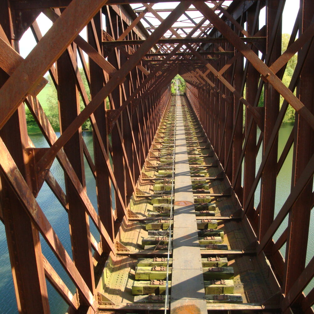 Blick auf eine alte, rostige Eisenbahnbrücke mit sichtbaren Stahlträgern und einem schmalen Weg in der Mitte, der über Wasser führt.