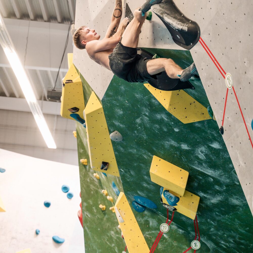 Ein Kletterer in kurzer Sportbekleidung klettert an einer vertikalen Kletterwand mit grüner und grauer Oberfläche. Gelbe und blaue Griffe sind sichtbar, sowie rote Sicherungsseile.
