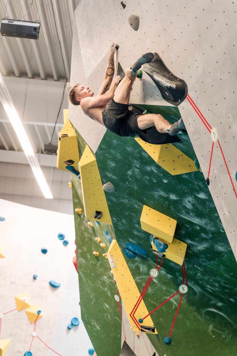 Ein Kletterer in kurzer Sportbekleidung klettert an einer vertikalen Kletterwand mit grüner und grauer Oberfläche. Gelbe und blaue Griffe sind sichtbar, sowie rote Sicherungsseile.