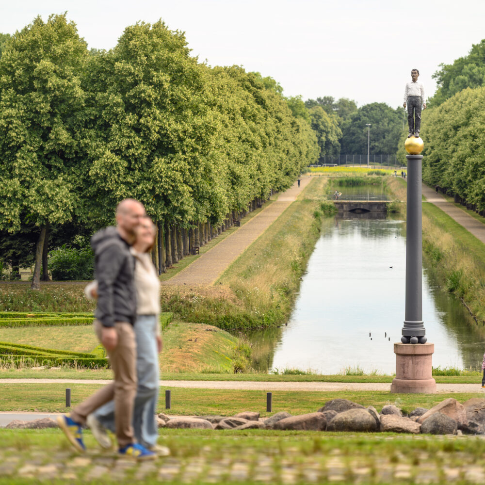 Pärchenläuft am Eisernen Mann vorbei und schaut auf die Gartenanlagen mit einem Wasserlauf und Bäumen im Hintergrund. Der Mann hat eine Glatze und trägt ein graues T-Shirt, die Frau hat kurze Haare und trägt eine weiße Bluse.