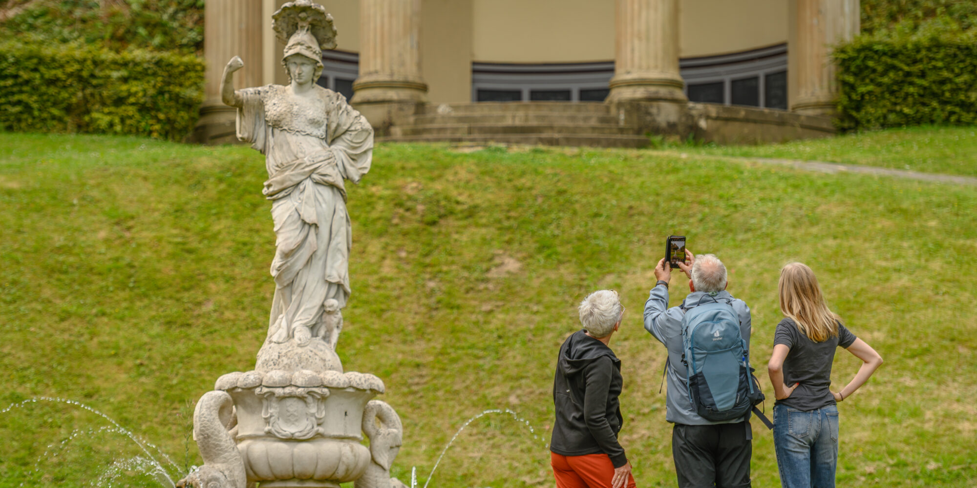 Eine Statue einer weiblichen Figur mit einer Schale auf dem Kopf steht auf einem Brunnen. Im Hintergrund sind drei Personen zu sehen, die ein Foto in den Gartenanlagen machen.