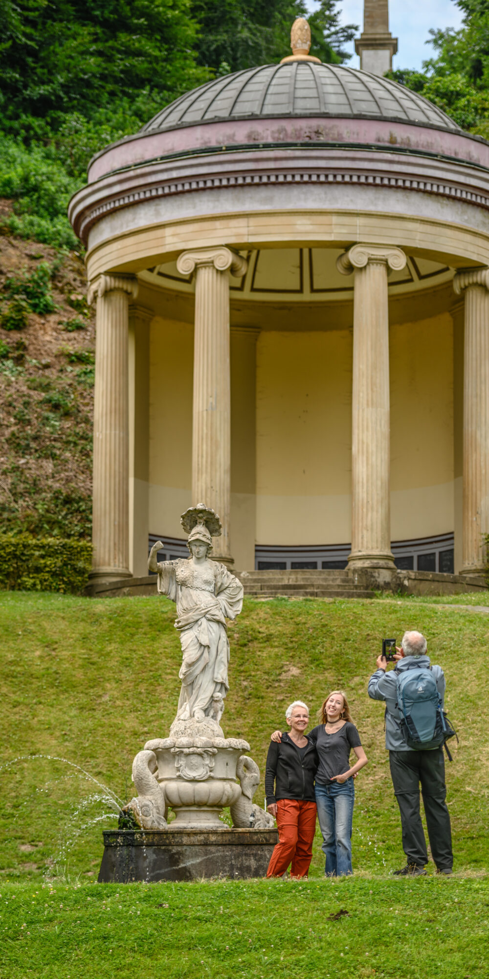 Runder Pavillon in den Gartenanlagen mit Kuppeldach und vier Säulen auf einem Hügel, davor eine steinerne Statue mit Wasserspielen und drei Personen, von denen eine fotografiert.