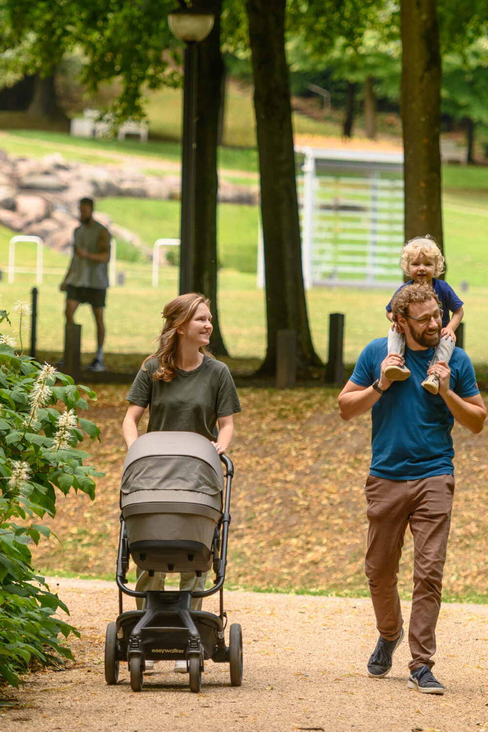 Eine Frau schiebt einen Kinderwagen auf einem Weg, während ein Mann ein Kind auf den Schultern trägt. Im Hintergrund ist eine Person zu sehen, die in einem Park steht.