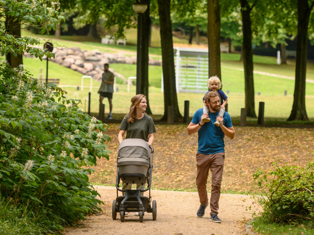 Eine Frau schiebt einen Kinderwagen auf einem Weg, während ein Mann ein Kind auf den Schultern trägt. Im Hintergrund ist eine Person zu sehen, die in einem Park steht.