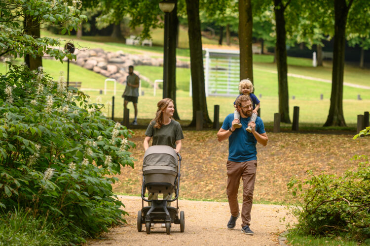 Eine Frau schiebt einen Kinderwagen auf einem Weg, während ein Mann ein Kind auf den Schultern trägt. Im Hintergrund ist eine Person zu sehen, die in einem Park steht.