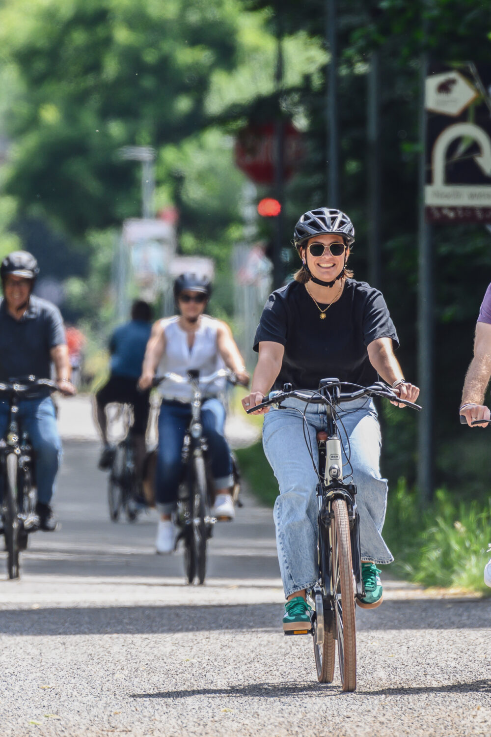 Radfahren in Kleve. Fünf Personen auf Fahrrädern fahren auf einem Radweg. Vier Personen tragen Helme, eine Person fährt ein E-Bike. Verkehrsschilder sind im Hintergrund sichtbar.