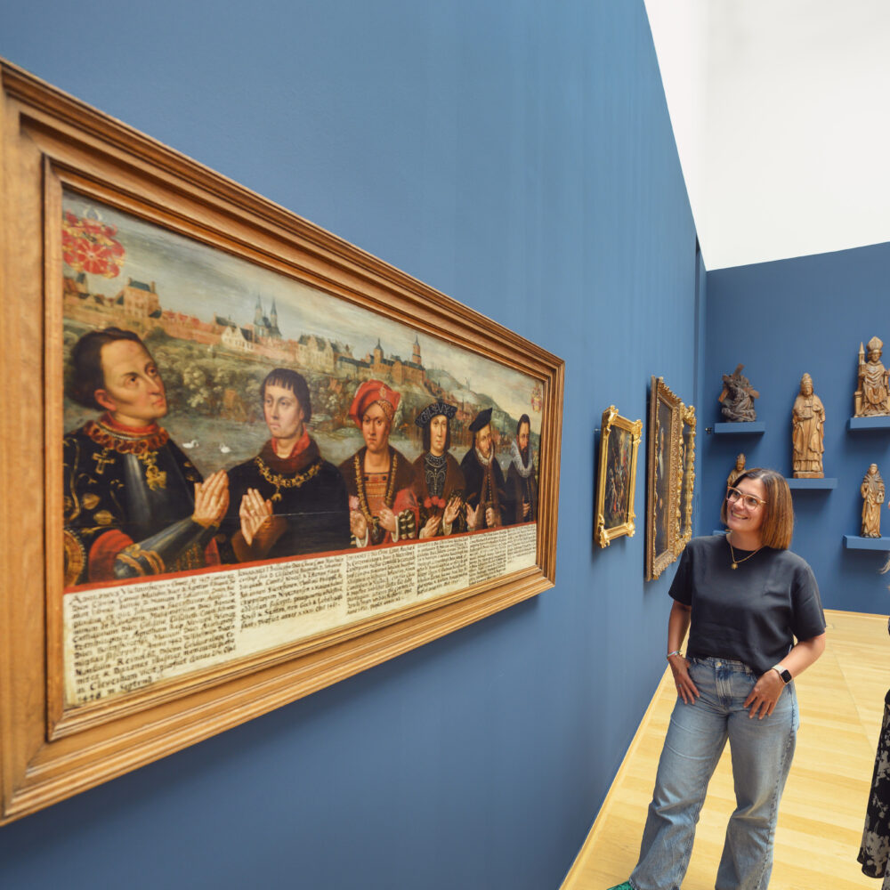 Zwei Personen stehen vor einem großen Gemälde mit historischen Figuren und einer beschrifteten Unterzeile. Im Hintergrund sind Holzskulpturen an der Wand zu sehen.