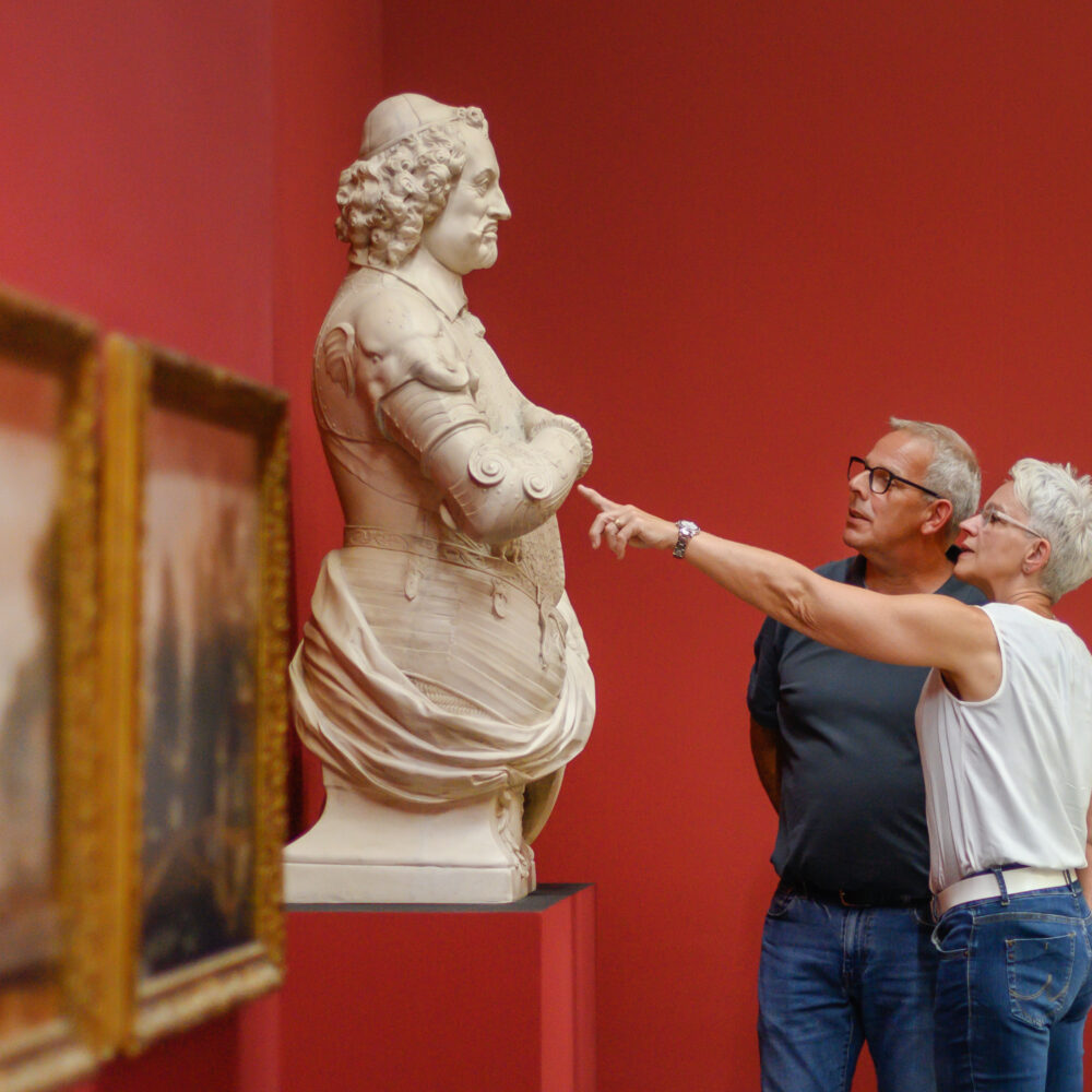 Zwei Personen stehen vor einer Skulptur in einem Museum. Die Skulptur zeigt eine weibliche Figur mit einem runden Kopf und einer detaillierten Kleidung. Im Hintergrund sind Bilderrahmen mit Gemälden an der Wand.
