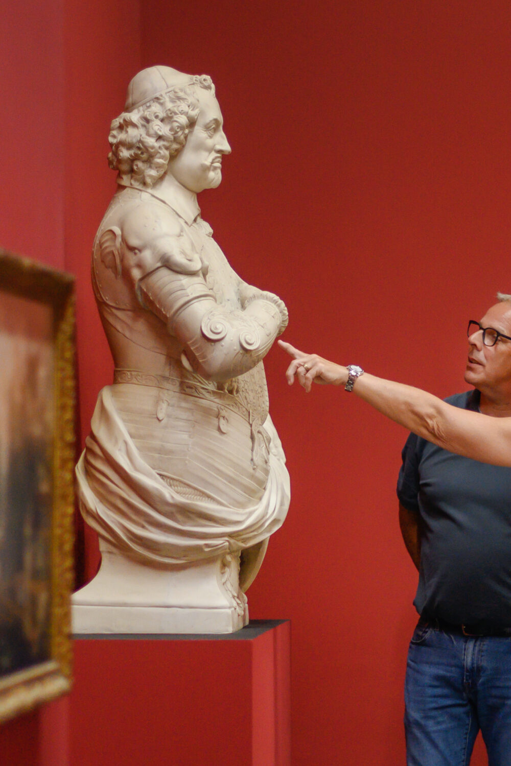 Zwei Personen stehen vor einer Skulptur in einem Museum. Die Skulptur zeigt eine weibliche Figur mit einem runden Kopf und einer detaillierten Kleidung. Im Hintergrund sind Bilderrahmen mit Gemälden an der Wand.