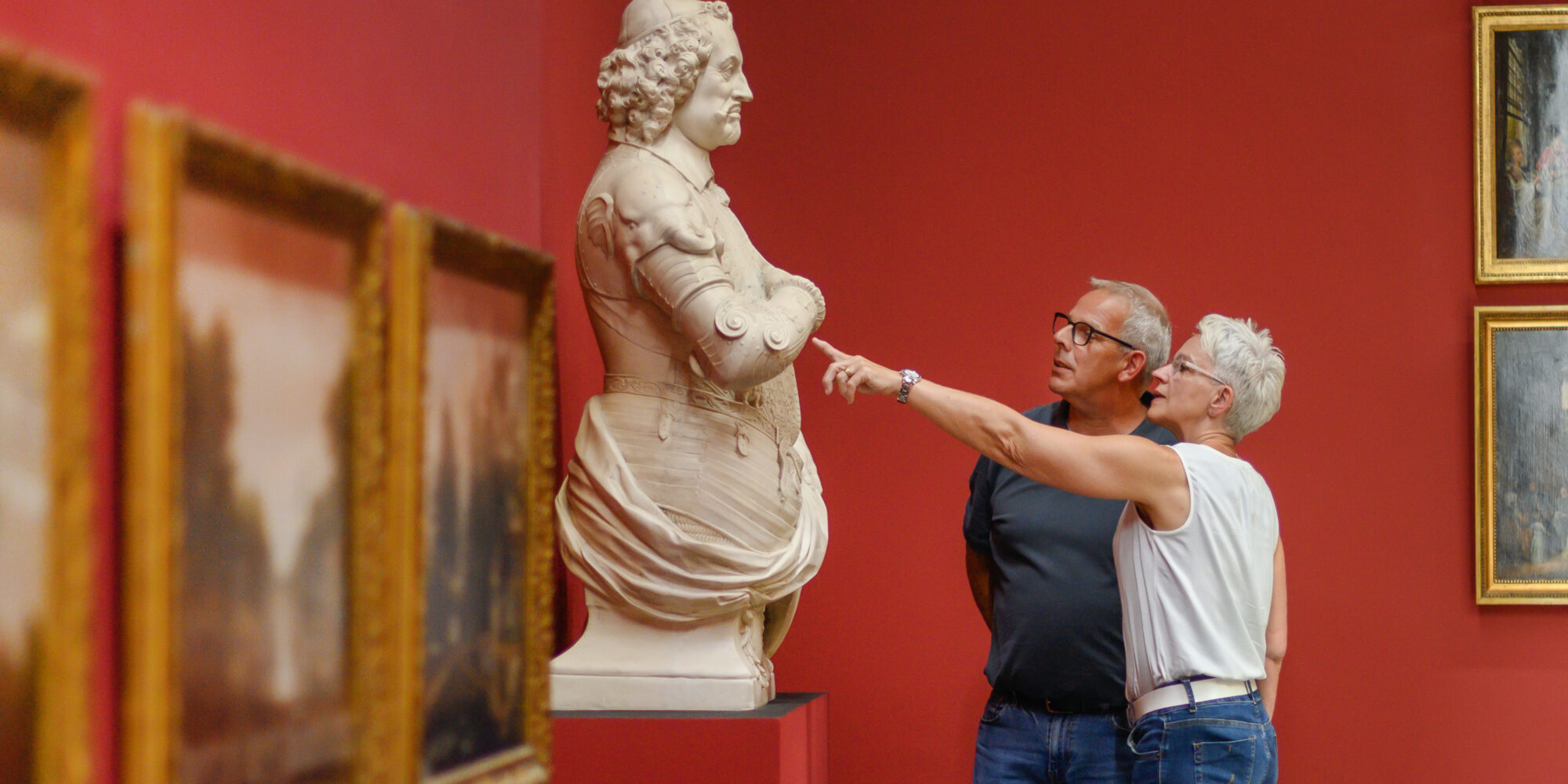 Zwei Personen stehen vor einer Skulptur in einem Museum. Die Skulptur zeigt eine weibliche Figur mit einem runden Kopf und einer detaillierten Kleidung. Im Hintergrund sind Bilderrahmen mit Gemälden an der Wand.