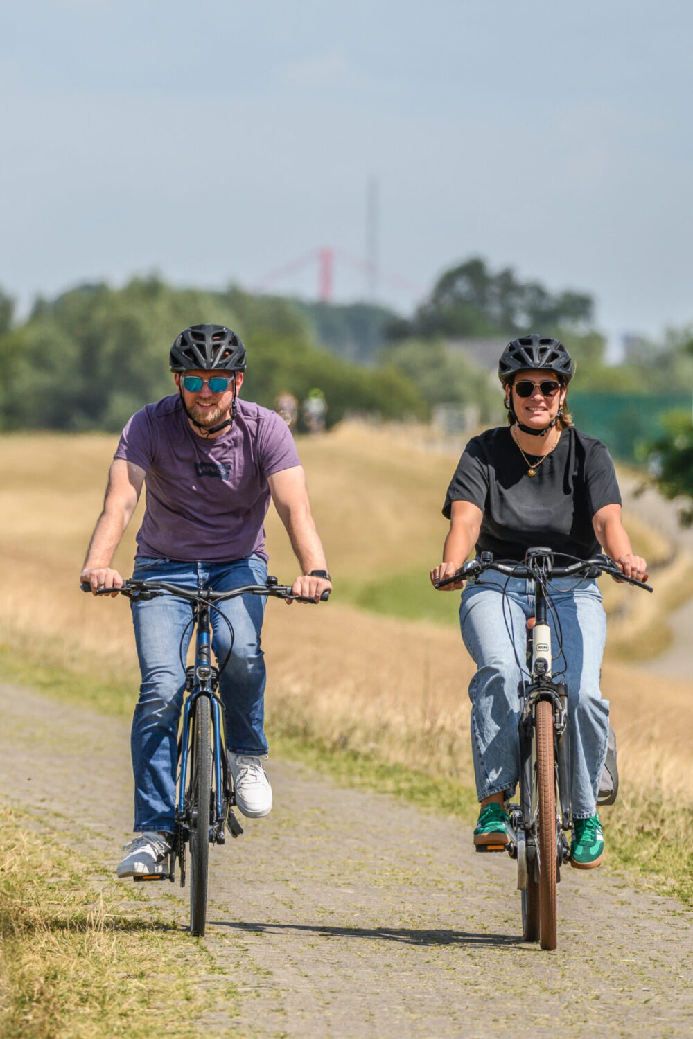 Zwei Personen auf Fahrrädern fahren auf dem Deich entlang einer Wiese. Im Hintergrund sind Bäume und der Rhein zu sehen.