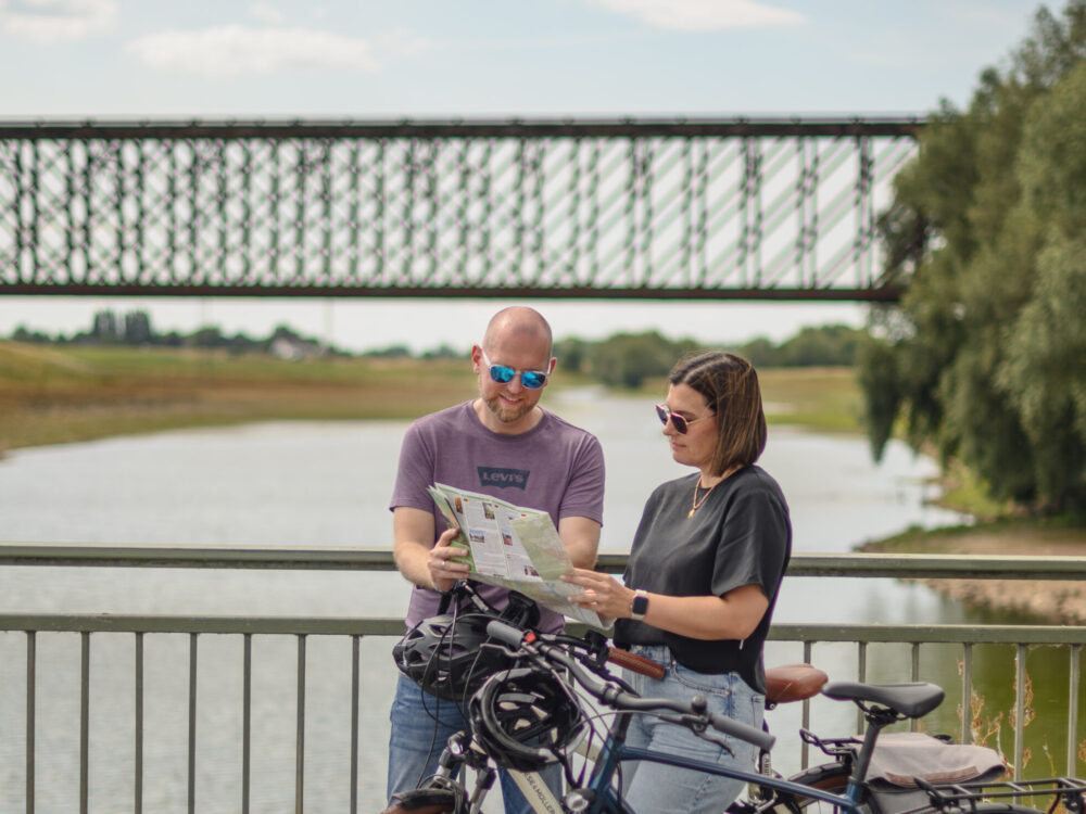 Ein Mann und eine Frau stehen an einem Geländer und betrachten eine Karte. Im Hintergrund ist eine Brücke über einen Fluss zu sehen. Zwei Fahrräder stehen neben ihnen.