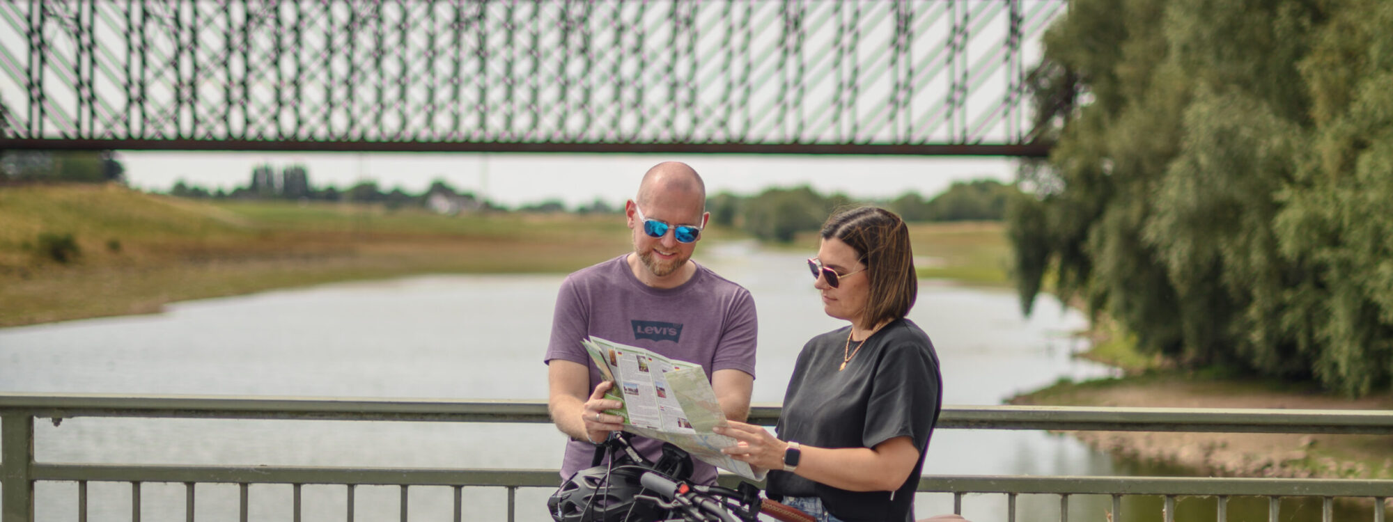 Ein Mann und eine Frau stehen an einem Geländer und betrachten eine Karte. Im Hintergrund ist eine Brücke über einen Fluss zu sehen. Zwei Fahrräder stehen neben ihnen.