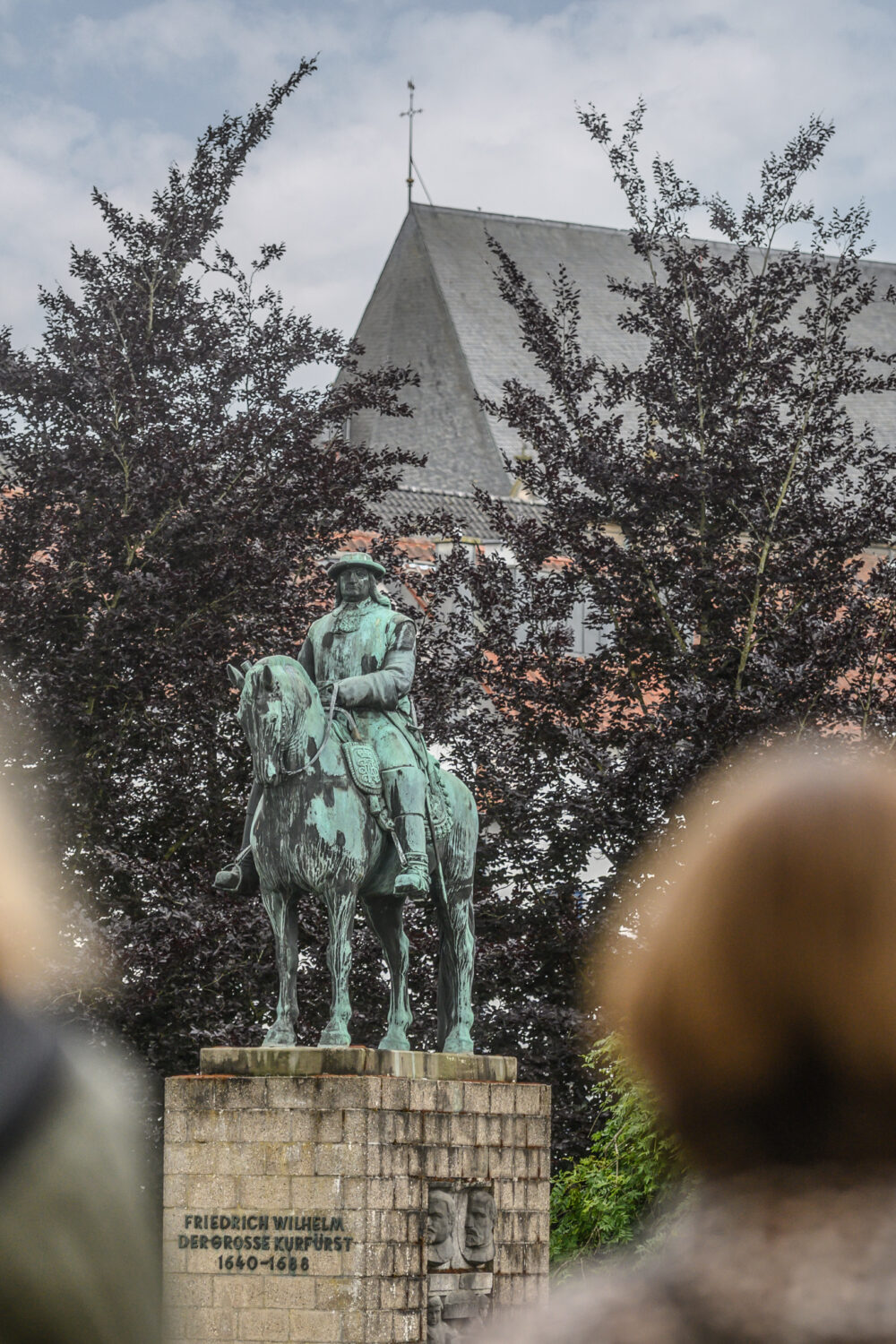 Der Große Kurfürst Friedrich Wilhelm als Bronzestatue auf einem Steinsockel. Zwei Frauen blicken auf das Denkmal des Großen Kurfürsten. Bäume und andere Gebäude sind im Hintergrund.