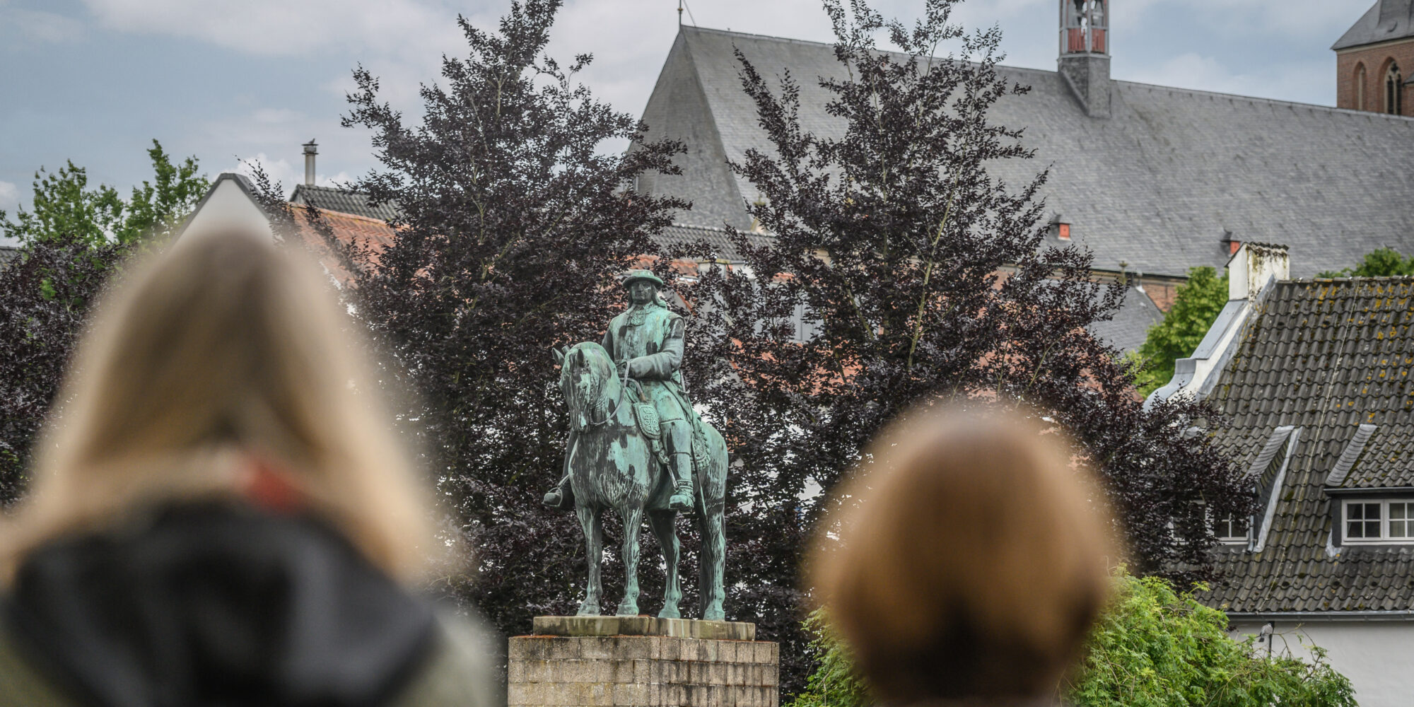 Der Große Kurfürst Friedrich Wilhelm als Bronzestatue auf einem Steinsockel. Zwei Frauen blicken auf das Denkmal des Großen Kurfürsten. Bäume und andere Gebäude sind im Hintergrund.