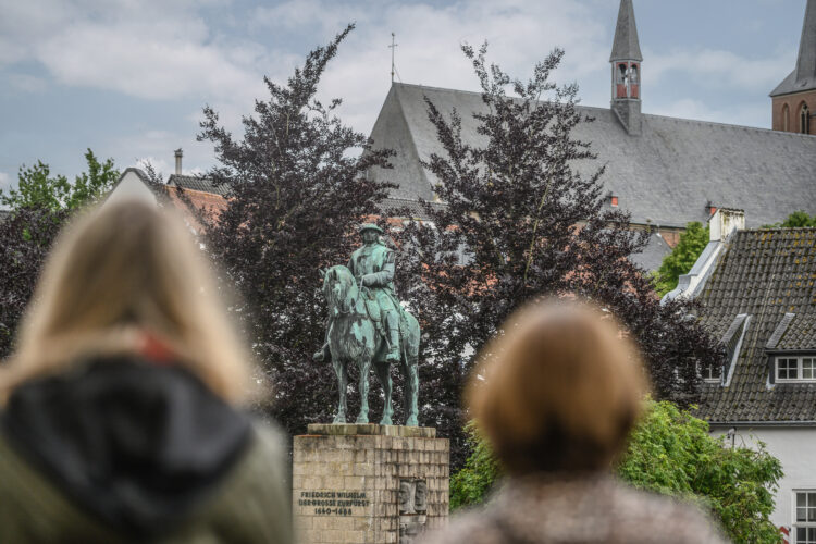 Der Große Kurfürst Friedrich Wilhelm als Bronzestatue auf einem Steinsockel. Zwei Frauen blicken auf das Denkmal des Großen Kurfürsten. Bäume und andere Gebäude sind im Hintergrund.
