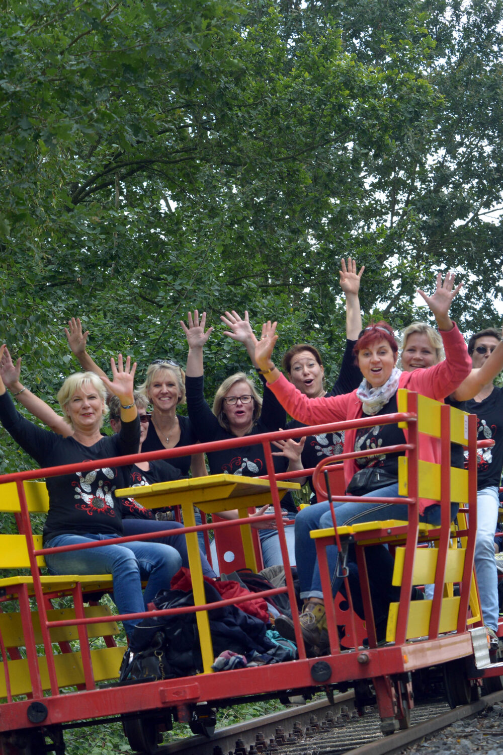 Mehrere Personen sitzen auf zwei offenen, roten und gelben Clubdraisinen und heben die Hände, umgeben von Bäumen und Gleisen.