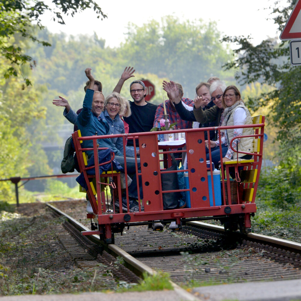 Eine rote Draisine mit mehreren Personen, die auf den Sitzen sitzen und die Hände heben. Im Hintergrund sind Bäume und ein Verkehrsschild mit der Aufschrift '100 m' sichtbar.