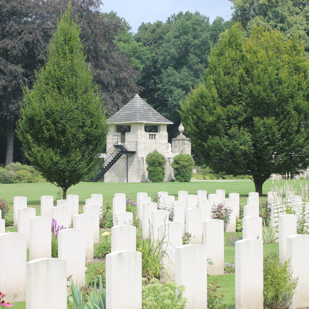 Weiße Grabmale auf dem Britischen Ehrenfriedhof auf einer grünen Wiese, Bäume im Hintergrund.