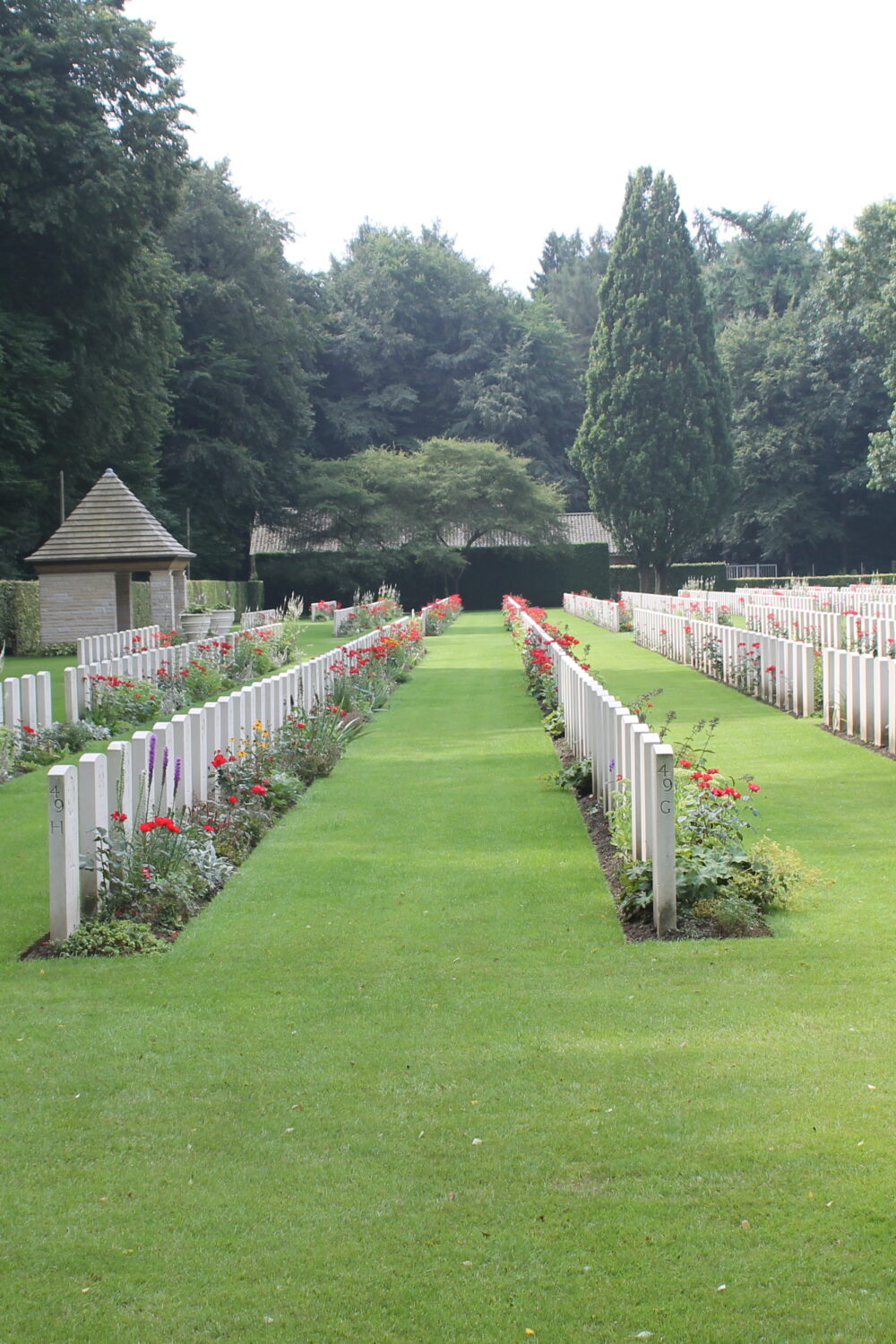 Weiße Grabmale auf dem Britischen Ehrenfriedhof auf einer grünen Wiese, Bäume im Hintergrund