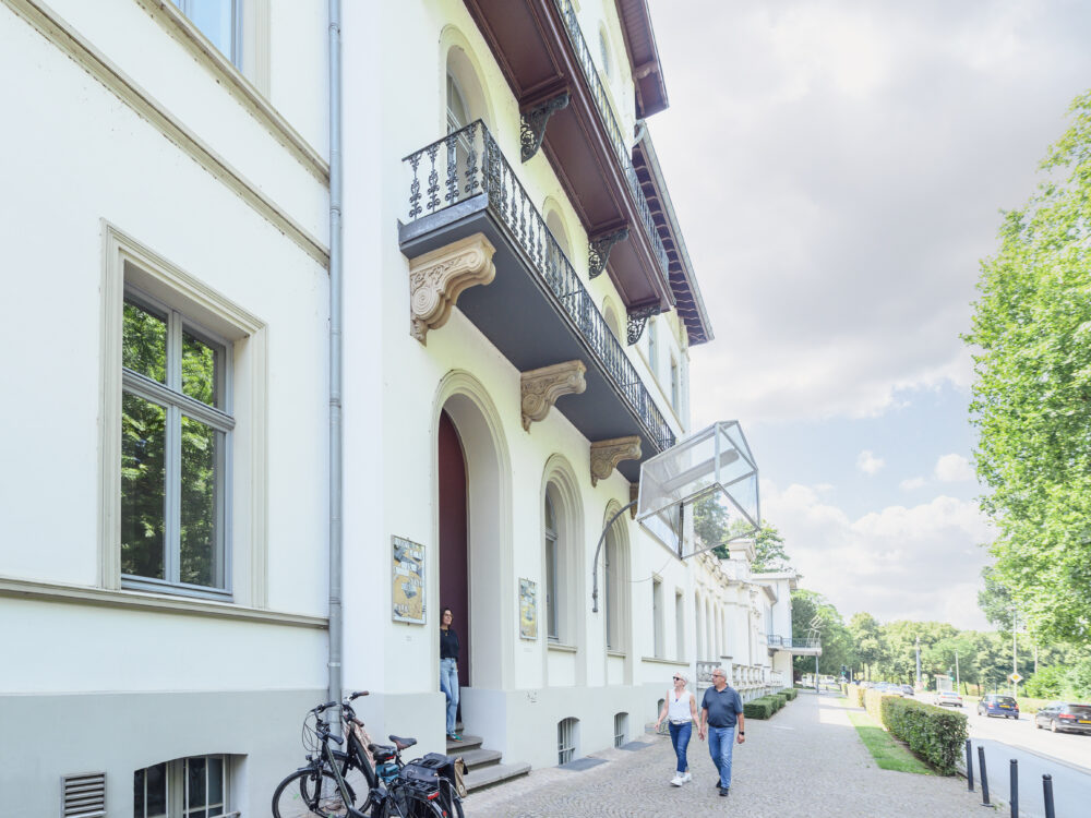 Fassade des Kurhauses in Kleve mit zwei Personen, die auf dem Gehweg gehen. Links stehen zwei Fahrräder an einem Fahrradständer.