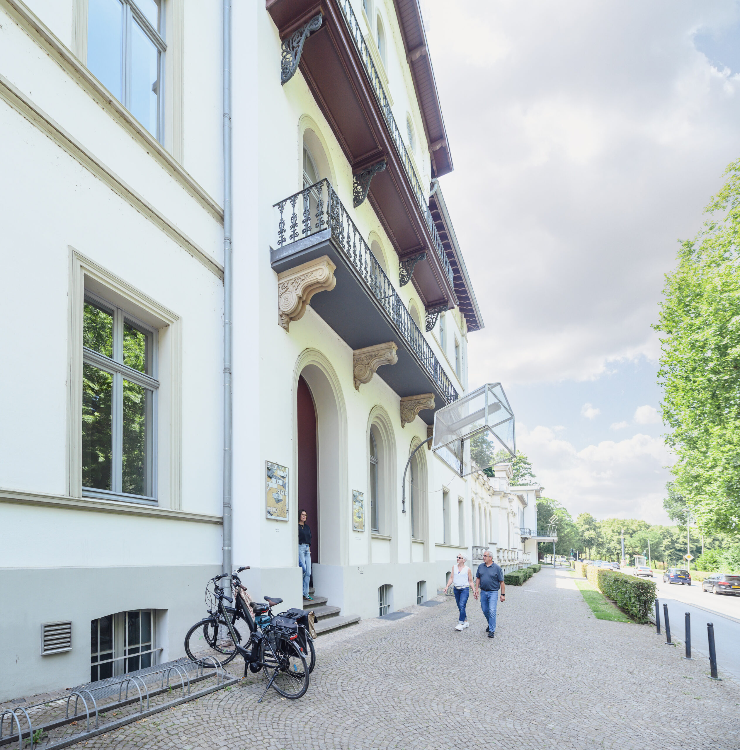 Fassade des Kurhauses in Kleve mit zwei Personen, die auf dem Gehweg gehen. Links stehen zwei Fahrräder an einem Fahrradständer.